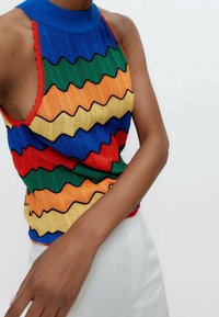Mouwloze gebreide top met meerkleurige zigzagstrepen in blauw, groen, oranje, geel en rood, gedragen met witte broek door een persoon.