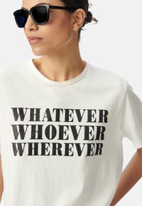 T-shirt blanc avec un texte noir pailleté indiquant "PEU IMPORTE QUI, PEU IMPORTE OÙ". Le modèle porte des lunettes de soleil noires. Manches courtes, col rond.