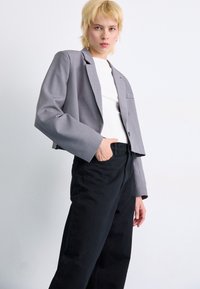 Gray cropped blazer with long sleeves and a single button, paired with black loose-fitting trousers, set against a light background.