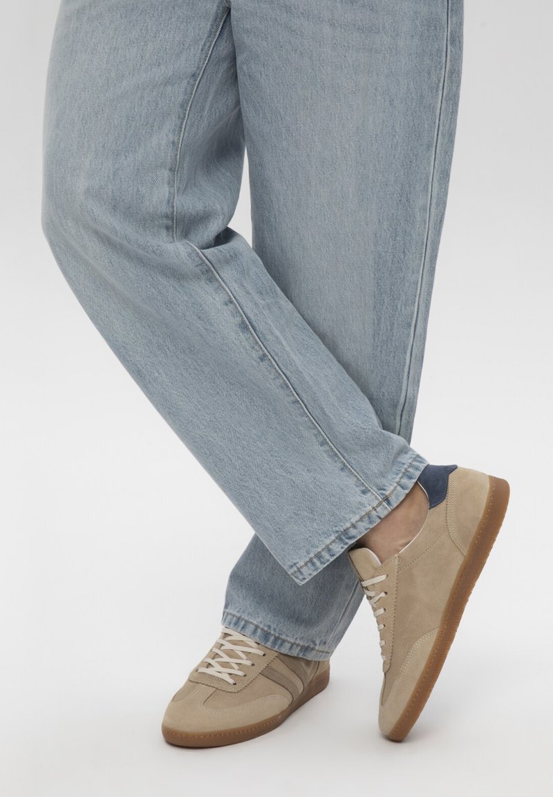 Personne portant un jean bleu clair et des baskets en daim beige avec des semelles en gomme, un pied croisé sur l’autre sur un fond uni.