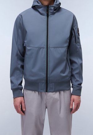 Uomo con giacca grigia con cappuccio e zip, con polsini e orlo a coste, abbinata a pantaloni beige chiaro, in piedi davanti a uno sfondo neutro.