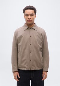 Νεαρός άνδρας που φορεί καφέ σακάκι με κουμπιά, γιακά και πλαϊνές τσέπες, στέκεται μπροστά σε απλό ανοιχτό γκρι φόντο.