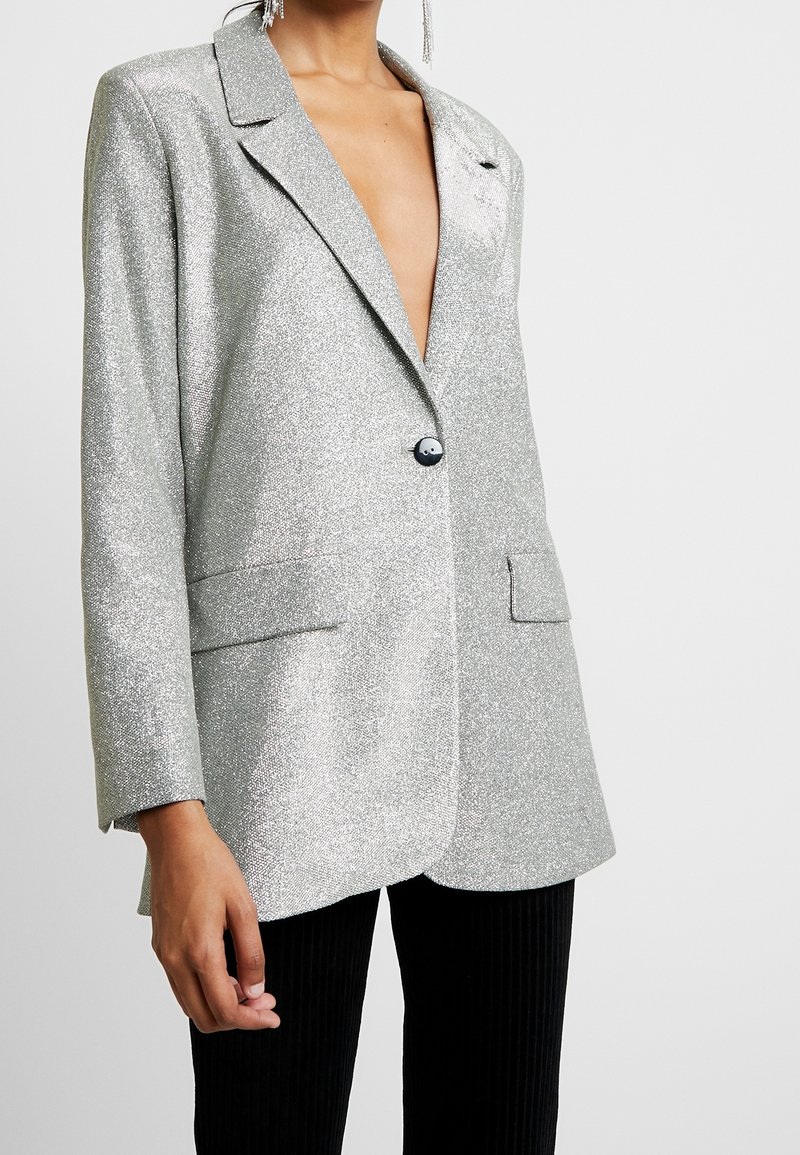 Femme portant un blazer pailleté argenté avec un seul bouton noir et un pantalon noir, montrée du cou aux hanches sur un fond uni.