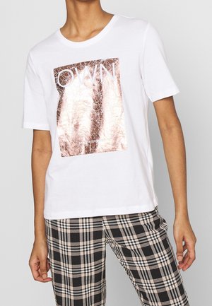 T-shirt blanc à manches courtes avec un carré doré rose brillant craquelé et le mot "OWN", porté avec un pantalon à carreaux noir et beige.