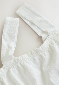 Vêtement en tissu blanc texturé avec larges bretelles et bord supérieur élastiqué et froncé.