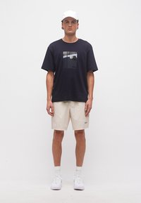 Νεαρός άνδρας που φοράει λευκό καπέλο, μαύρο γραφικό μπλουζάκι, μπεζ σορτς, λευκές κάλτσες και λευκά αθλητικά παπούτσια, στέκεται μπροστά σε απλό λευκό φόντο.