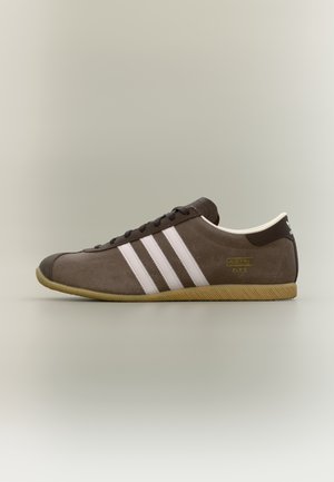 Matala profiili ruskea mokkakenkä, jossa valkoiset kolme raitaa, kumipohja ja "Adidas Paris"-brändäys neutraalilla taustalla.