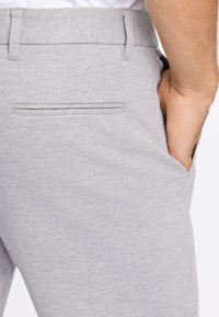 Pantalon en textile gris avec une taille plate, poches latérales et une poche arrière avec un détail en piping. Le tissu semble doux et texturé.
