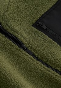 Fleecejacke in olivgrün mit strukturierter Oberfläche. Verfügt über einen schwarzen Reißverschluss und eine Reißverschlusstasche auf der Vorderseite.