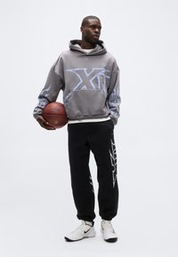 Mann in grauem Kapuzenpullover und schwarzen Jogginghosen hält einen Basketball, steht vor einem schlichten hellen Hintergrund und trägt weiße Nike-Sneaker.