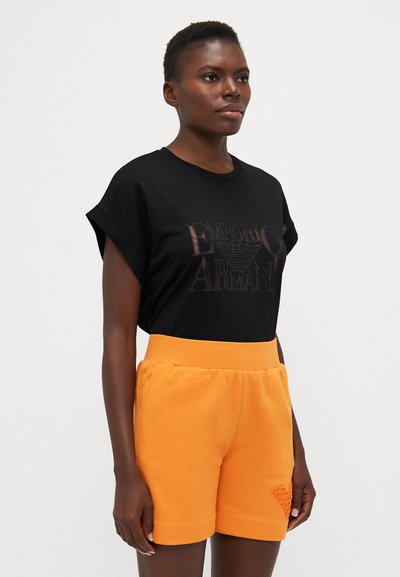 Black cotton t-shirt with rolled sleeves and a chest logo, paired with orange fleece shorts featuring a textured pocket detail.