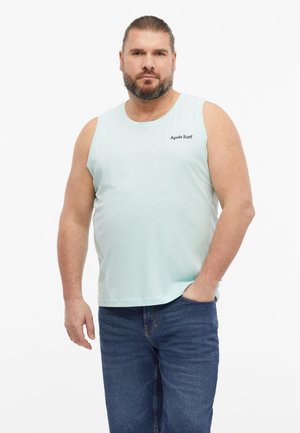 Mężczyzna z brodą, noszący jasnoniebieski bezrękawnik z napisem "Après Surf" oraz niebieskie dżinsy, stoi z jedną ręką w kieszeni.