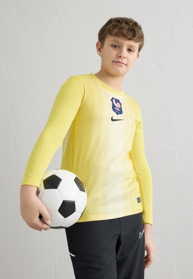 Hosszú ujjú sárga futballmez függőleges csíkokkal, kék logóval és fekete Nike logóval, fekete rövidnadrággal és egy futballlabdával párosítva.