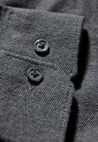 Donkerdenimstof met een gestructureerd oppervlak, voorzien van twee zwarte knopen op een opgerolde kraag. De knopen hebben elk vier gaatjes.