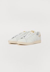 Sapatilhas de couro brancas com um design de cano baixo, detalhes perfurados nas laterais e uma aba no calcanhar em bege com um logótipo dourado. Sola de borracha.