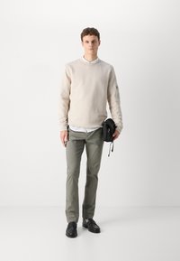 Lys beige sweatshirt, slanke olivengrønne bukser, sorte sko. Modellen holder en lille sort taske, der har en sidelomme og lynlåsdetailler.