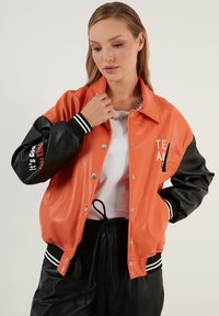 Oranje en zwarte gewatteerde jas met drukknoopsluiting, geribbelde manchetten en vetgedrukte tekst op de mouw. Kenmerkt een glanzende uitstraling en een oversized pasvorm.
