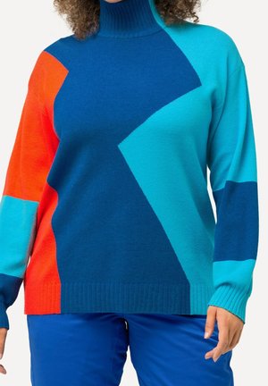 Woman wearing a color-blocked turtleneck sweater in orange, dark blue, and turquoise with matching blue pants against a white background.