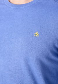 Helles blaues Strickpullover mit rundem Halsausschnitt und kleinem gelben "&"-Logo auf der linken Brustseite. Glatte Textur.