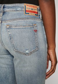 Light blue denim jeans with a textured surface, featuring a brand patch, yellow and orange stitching, and a small red accent on the back pocket.