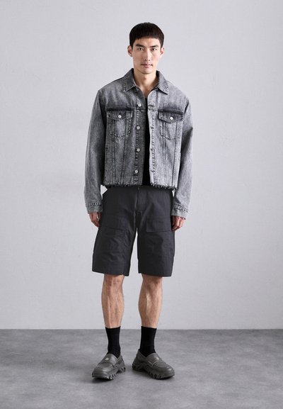 Veste en denim gris avec deux poches poitrine, t-shirt noir en dessous, shorts cargo noirs, chaussettes noires et chaussures slip-on grises avec semelle texturée.