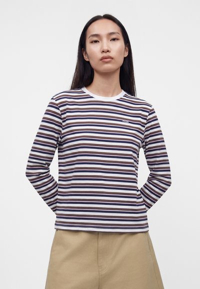 Γυναίκα που φοράει λευκό, ναυτικό μπλε και καφέ οριζόντιες ρίγες μακρυμάνικο πουκάμισο με μικρό λογότυπο στο στήθος και μπεζ παντελόνι, στέκεται μπροστά σε απλό φόντο.