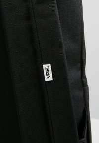 Zaino in tela nera con cuciture verticali, dotato di un piccolo etichetta logo bianca. Materiale con finitura testurizzata. Tasca laterale visibile.