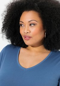 Tričkot s V-výstřihem a krátkým rukávem v měkké, texturované modré tkanině; má mírný lesk a volný střih, který zdůrazňuje hladký výstřih a tělo.