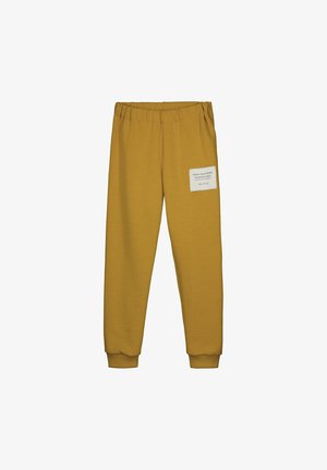 Pantalones jogger de color mostaza con cintura y puños elásticos, con un parche blanco con texto en el muslo derecho.