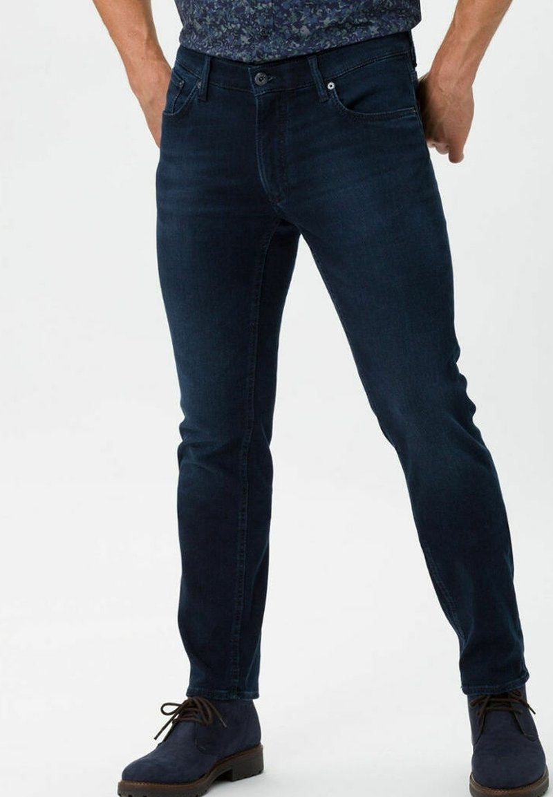 Man wearing dark blue slim-fit jeans and navy blue lace-up boots, standing against a plain light background.