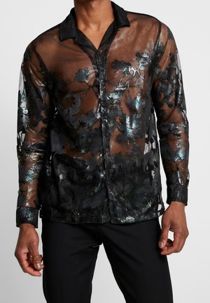 Man draagt een doorschijnend zwart overhemd met bloemmotief, glanzende metalen accenten en een zwarte broek tegen een effen achtergrond.