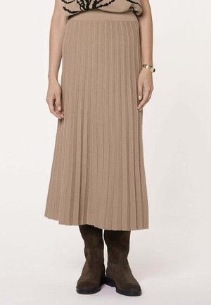 Beige geplooide midirok gedragen met bruine suède laarzen en een gouden armband aan de rechterpols, getoond van taille tot voeten.