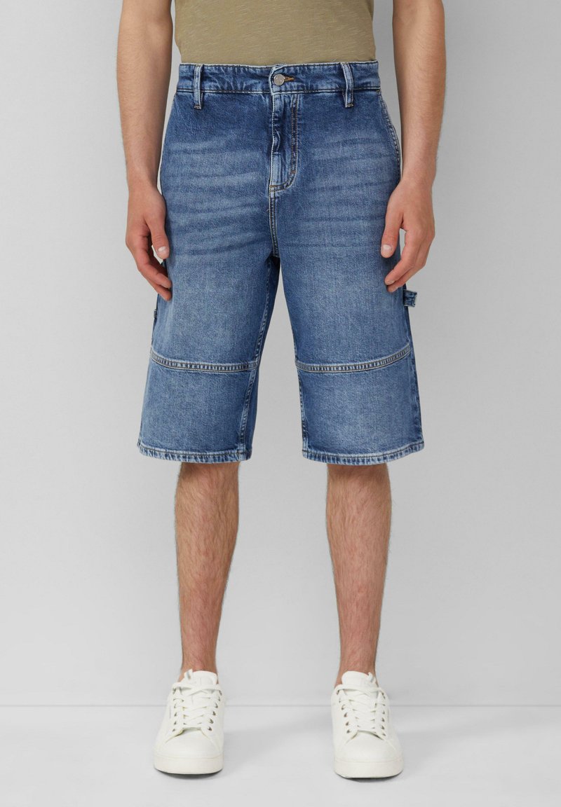 Shorts de mezclilla en un lavado azul medio, con un corte recto y largo hasta la rodilla, y costuras en contraste. Combinados con zapatillas blancas.