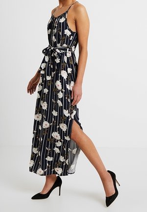 Kobieta w granatowej sukience bez rękawów z białym kwiatowym wzorem i pionowymi paskami, z bocznym rozcięciem, wiązaną w talii, nosząca czarne szpilki, idąca po białej podłodze.