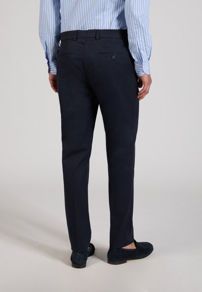 Uomo che indossa pantaloni eleganti blu navy, una camicia azzurra a righe e mocassini scuri, in piedi con la schiena visibile su uno sfondo semplice.