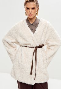 Faux fur frakke i hvid med en blød, struktureret overflade, åben front, brede ærmer og et brunt taljebælte. Har en v-udskæring.