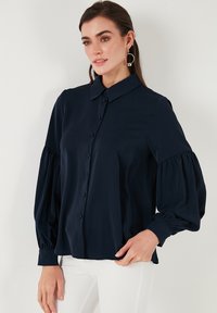 Camicia blu navy a bottoni realizzata in tessuto morbido, con colletto, maniche lunghe a sbuffo e polsini arricciati. Pantaloni bianchi visibili.