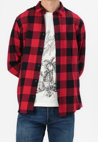 Hombre con una camisa de cuadros en rojo y negro, abierta sobre una camiseta gráfica blanca con palmeras y jeans azules.