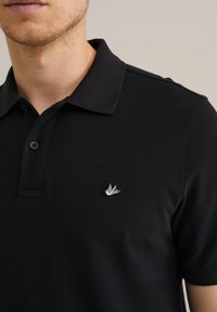 Melna polo krekla izstrādājums no teksturēta auduma, ar trīs pogām un nelielu baltu izšūtu logo krūtīs.