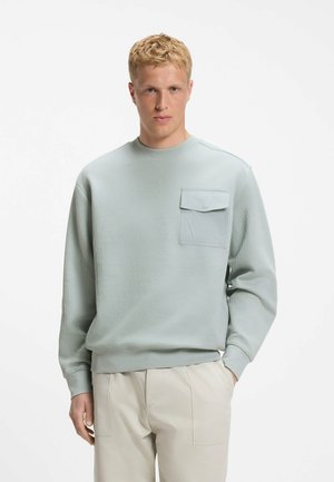 Blonder Mann trägt einen hellblauen Pullover mit langen Ärmeln und Brusttasche sowie beigen Hosen und steht vor einem schlichten weißen Hintergrund.