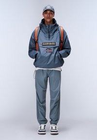 Water-resistant blue jacket with a hood and logo, paired with matching trousers. Features orange straps and white sneakers with navy accents.