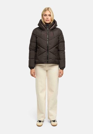 Femme portant une doudoune noire à capuche, un pantalon large crème et des baskets beiges, debout face à l’avant sur un fond uni.