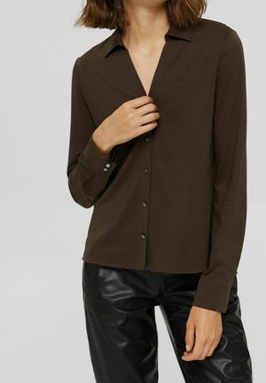 Kobieta ubrana w ciemnobrązową, zapinaną na guziki koszulę z kołnierzykiem oraz czarne skórzane spodnie, trzymająca koszulę jedną ręką przy kołnierzyku.