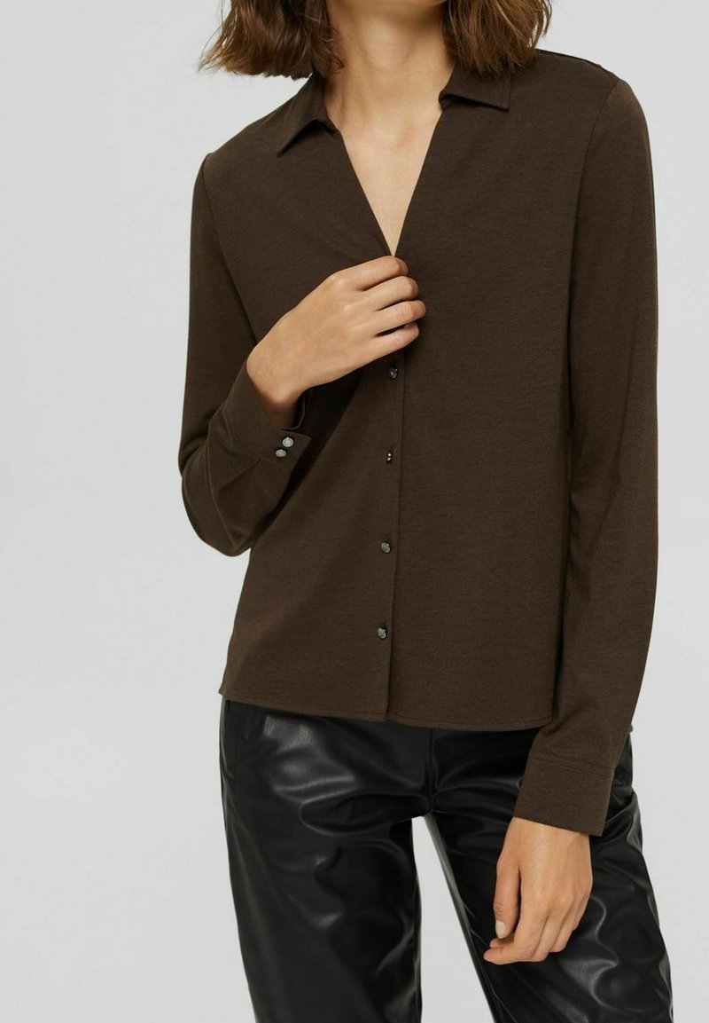 Femme portant une chemise marron foncé boutonnée avec un col et un pantalon en cuir noir, tenant la chemise près du col d'une main.