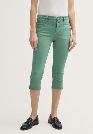 Femme portant un pantalon court vert avec taille à double bouton et des mocassins noirs, debout devant un fond clair uni.
