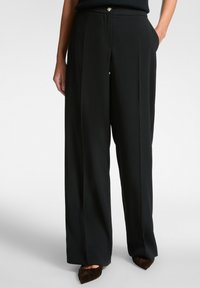 Femme portant un pantalon large noir avec des poches latérales et un top noir, associé à des chaussures noires à bouts pointus.