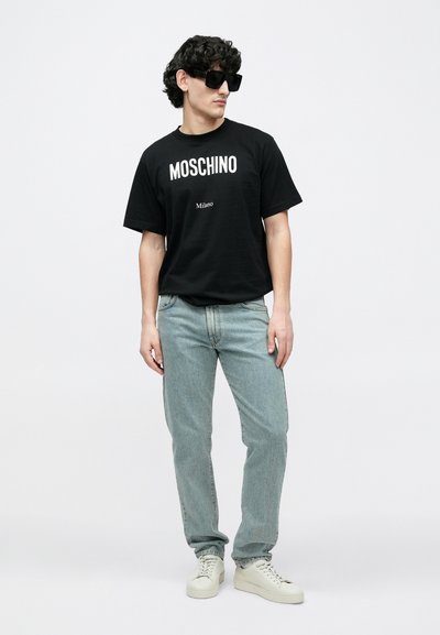 Juoda medvilninė marškinėliai su baltu užrašu „MOSCHINO“ ir „Milano“, derinami su šviesiai mėlynomis džinsais ir baltais sportbačiais, pasižymintys laisvu siluetu.