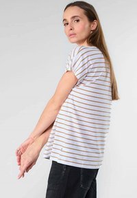 Camiseta blanca de manga corta con finas rayas marrones, corte holgado, tejido suave, dobladillo curvado, combinada con vaqueros oscuros.