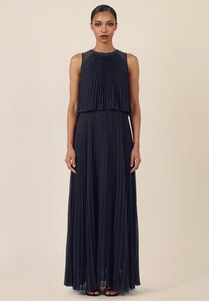 Frau mit glattem Haar, die ein ärmelloses, marineblaues, plissiertes Maxikleid und schwarze offene Schuhe trägt, steht vor einem schlichten beigen Hintergrund.