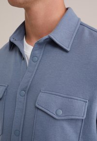 Košile na knoflíky z lehké modré látky, s límečkem, dvěma náprsními kapsami a kulatými knoflíky. Hladká textura s viditelnými detaily švů.